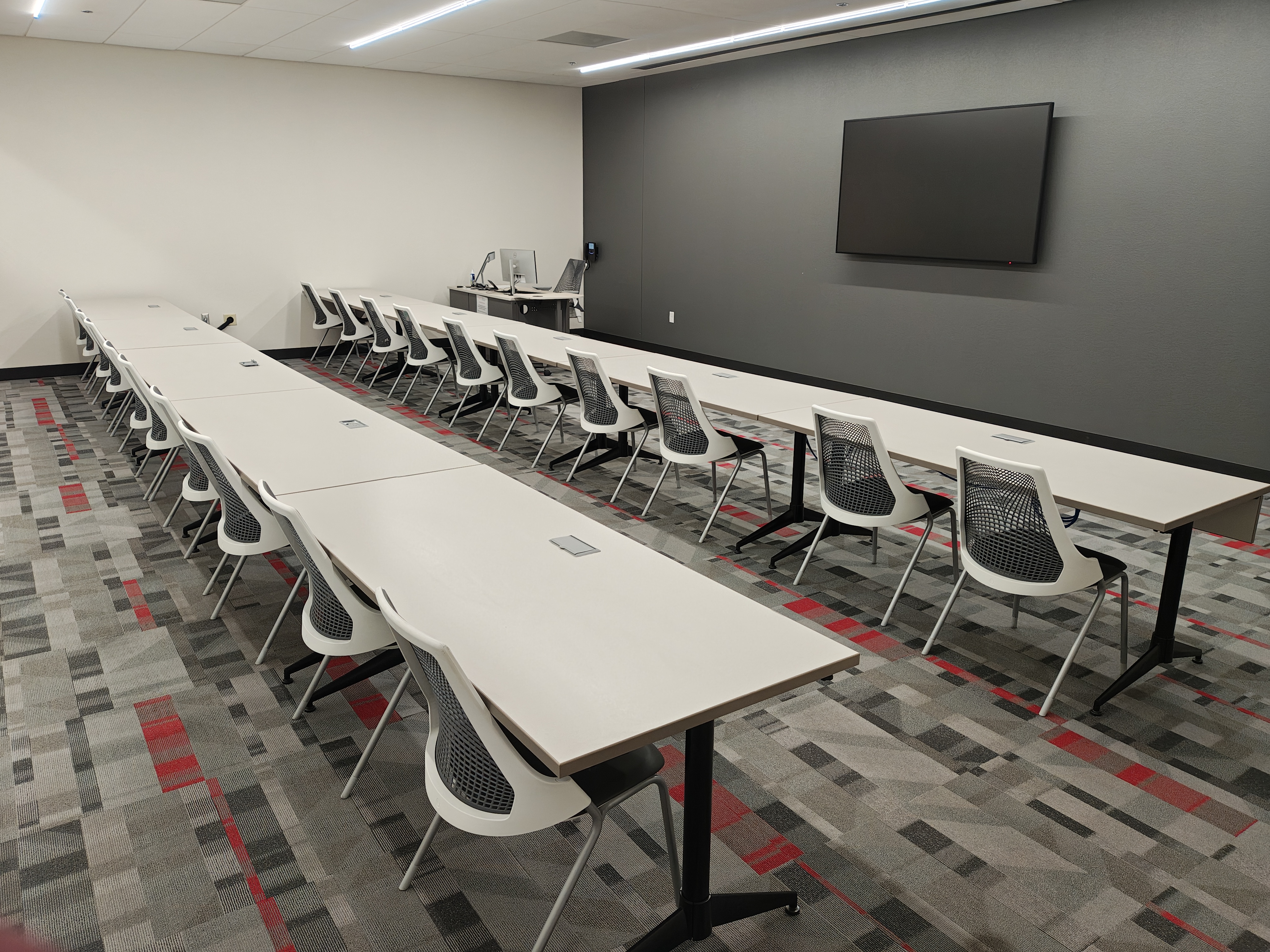 Empty classroom with rows of tables and chairs and a TV screen in front 